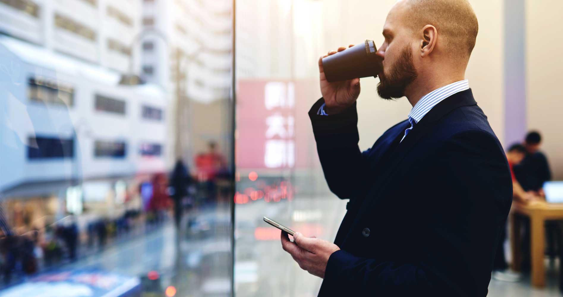 Man looking out of an office window on his mobile drinking a coffee